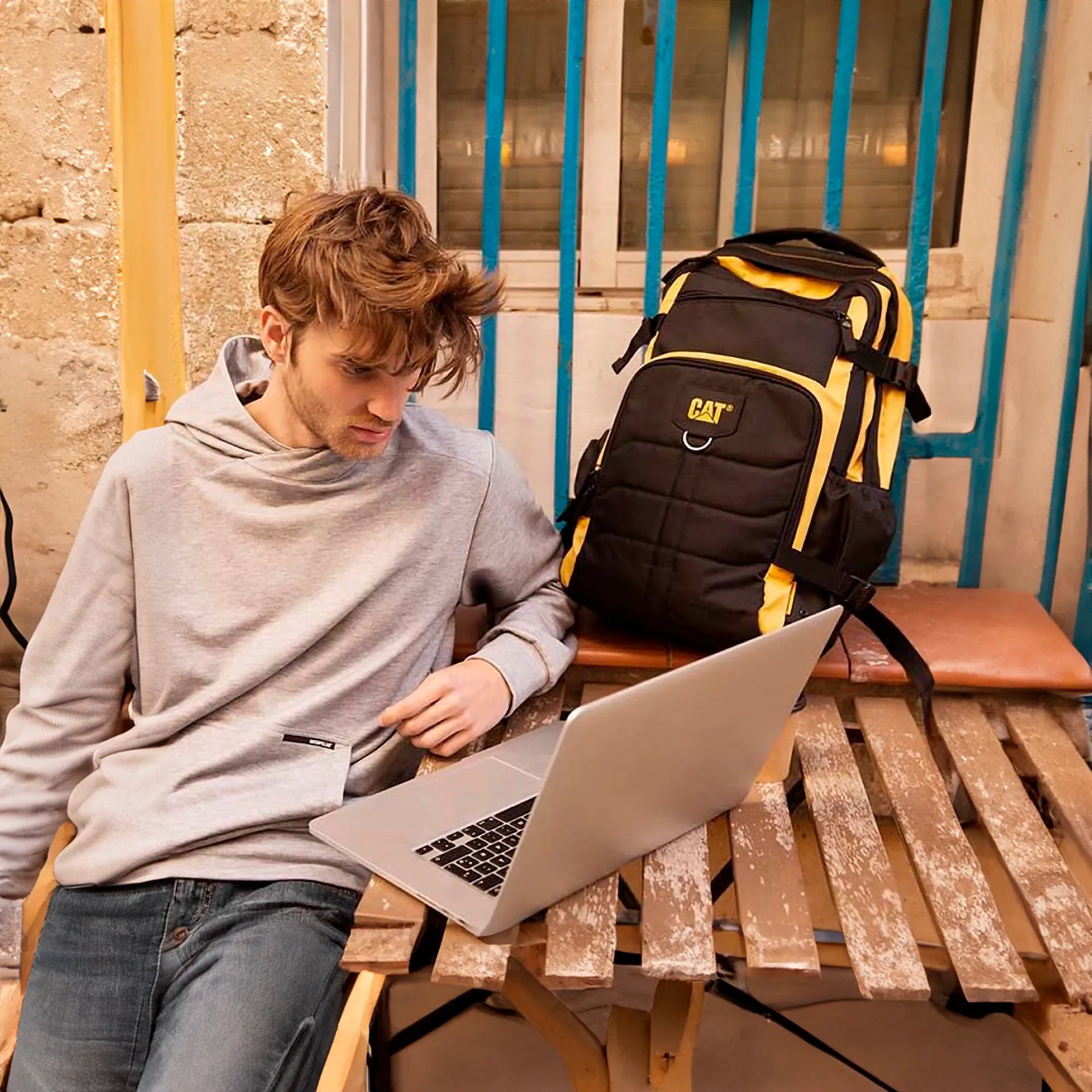Joven sentado usando una laptop con una mochila Caterpillar negra y amarilla.